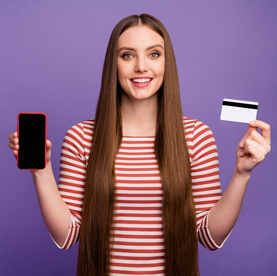 Positive girl hipster hold smartphone credit card present modern technology promotion she pay easy bank payment service wear striped white sweater jumper isolated purple color background